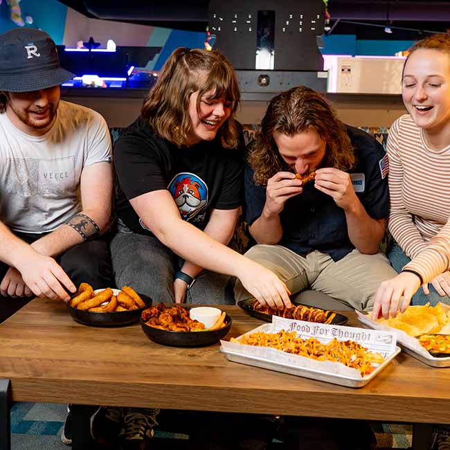 Four friends sit on a couch, laughing and sharing food, drinks, wings, and fries on a table.