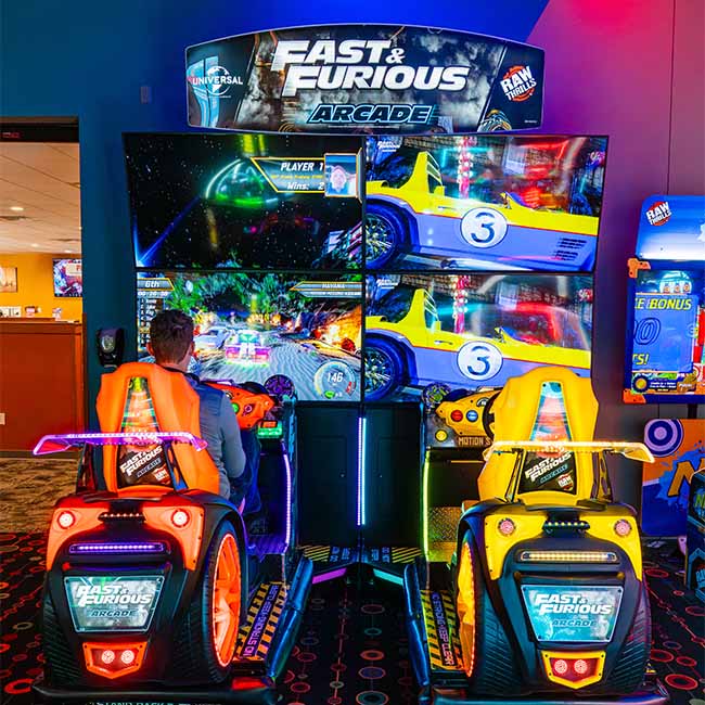 Two people play a Fast & Furious racing arcade game, enjoying food in a colorful game room.